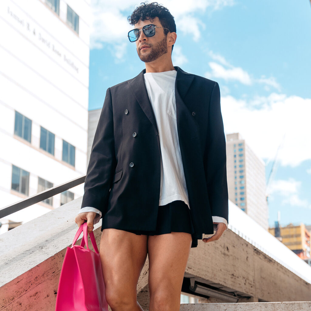 Gilbert Bolden III walks down a white marble stairway outdoors in New York City on a sunny day. He wears a black blazer, large white T-Shirt, and black mini-skirt. Wearing sunglasses, he looks down as he walks down the stairs, keeping his arms in close and carrying a hot pink bag in his right hand.