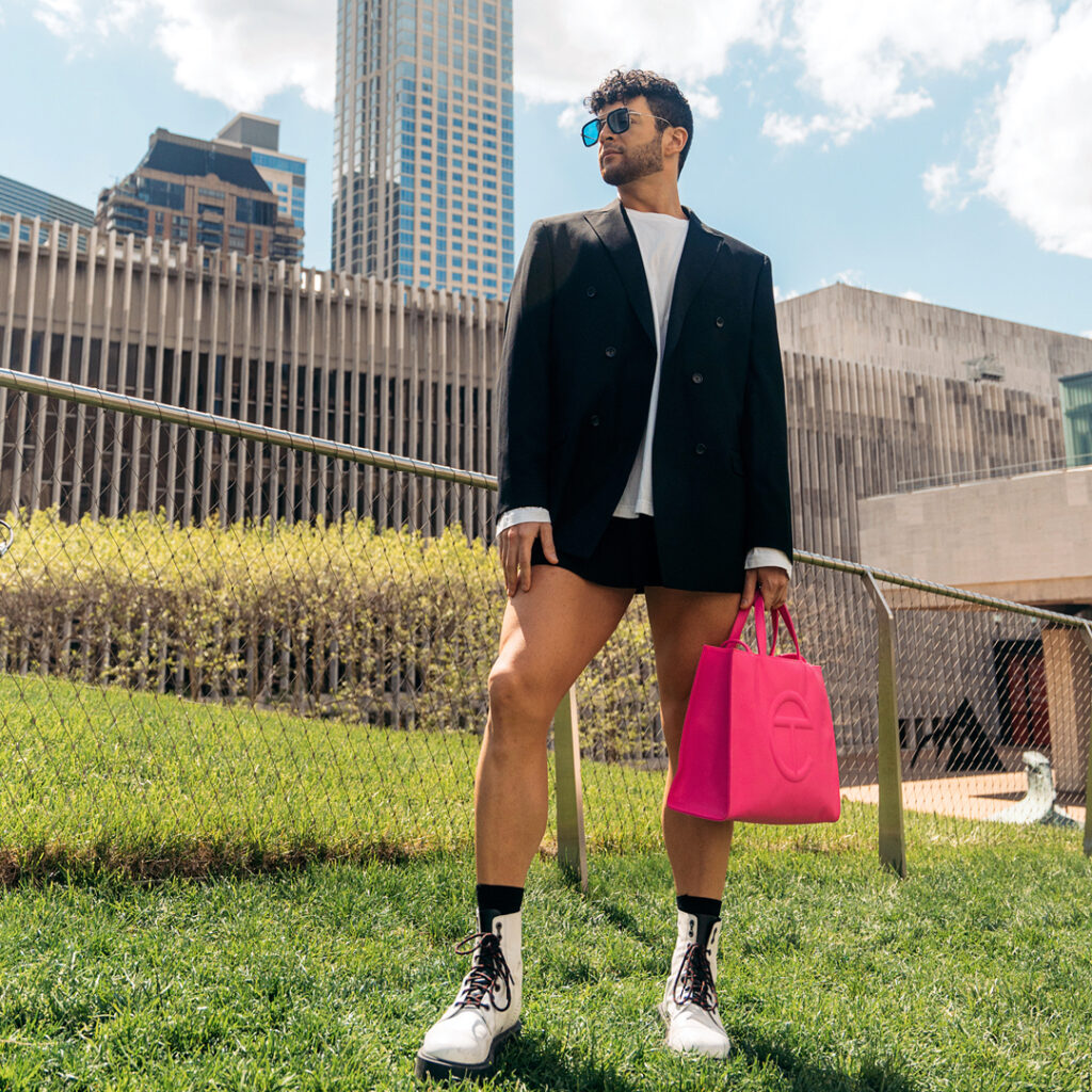 Gilbert Bolden III walks along a grassy knoll outdoors in New York City on a sunny day. He wears a black blazer, large white T-shirt, black mini-skirt, and high-top white boots with black laces and chunky soles. He looks over his right shoulder and carries a hot-pink bag in his left hans. A large high-rise building looms in the background.