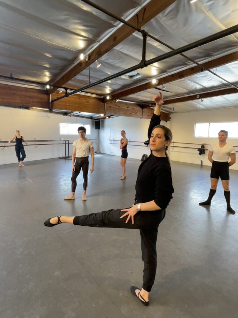 Melissa Barak leads a rehearsal at Los Angeles Ballet in a large studio with wood beams on the ceiling. She demonstrates a low developpe side.