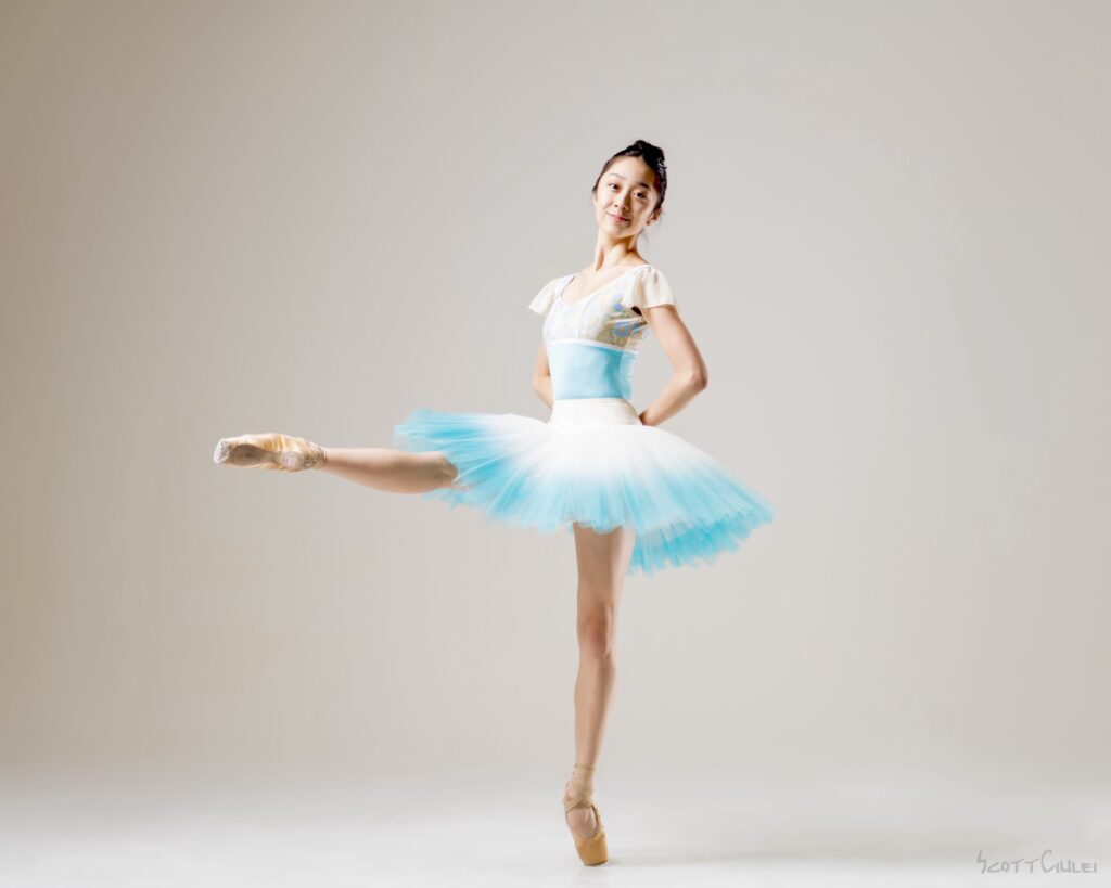 Sumi Ichikawa wears a white and teal ombré pancake tutu as she poses for a photo on pointe, her right leg extended in a battement devant. She rests her hands on her lower back and smiles.