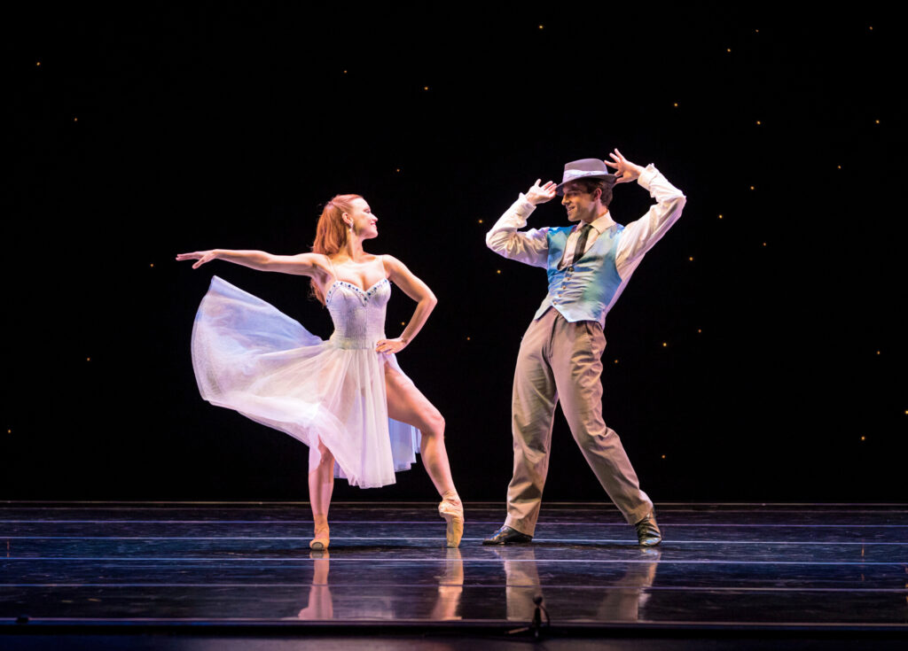 Erin Yarbrough-Powell and Robert Kretz face each other onstage in front of a starlit black backdrop. She wears a filmy white dress, and she tosses the skirt up while lifting her right arm to the side and holds her left hand on her hip, popping up her left foot onto pointe. Kretz, in a white shirt, light blue vest and khaki pants, tips his hat to her. They smile at each other coyly, clearly having fun.