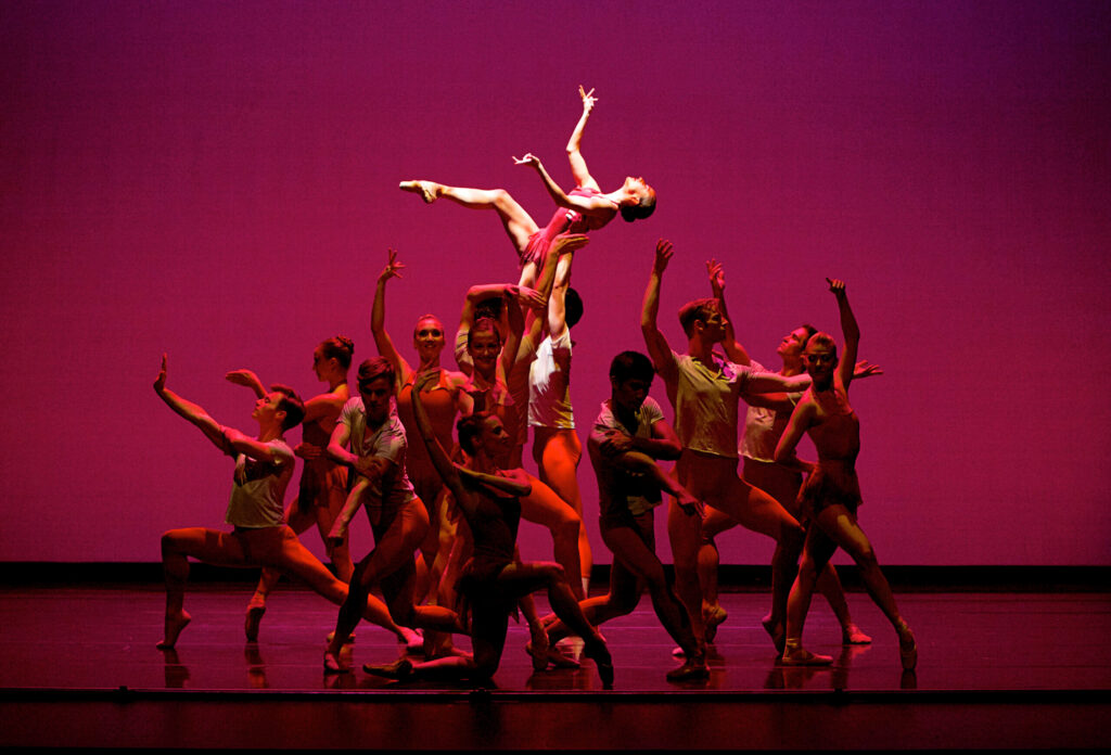 A large group of dancers creates a dramatic tableau onstage, with one spotlit woman lifted high above the group with her right leg lifted and bent and her arms out, hands splayed. The corps of dancers are bathed in a dark pink light and pose in front of a fuchsia backdrop.