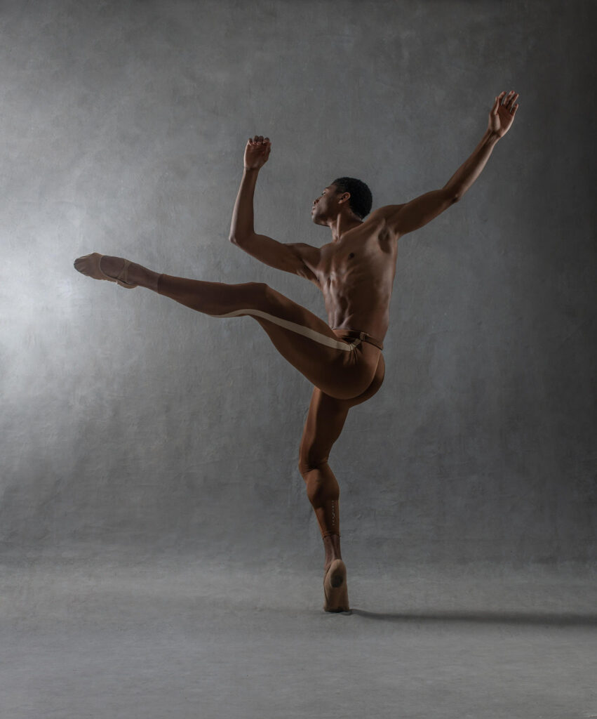 Nasrullah Abdur-Rahman poses in attitude devant, profile. He wears dark reddish-browns ballet tights and lifts his chest up as he raises his arms overhead.