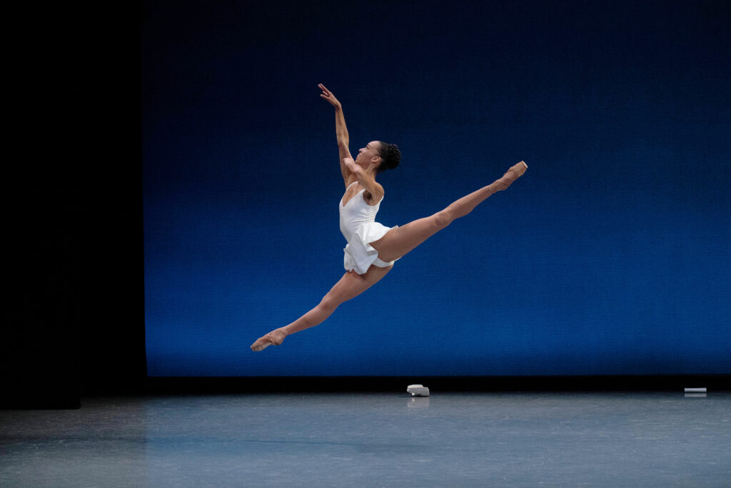 During a performance onstage in front of a blue backdrop, Chyrstyn Fentroy does a huge sissone fermé en avant, her legs split in the air. She holds the pointer finger of her left hand to her lips and raises her right arm up in front of her, and she looks up toward her hand and smiles. She wears a short white dance dress, brown tights, and brown pointe shoes.