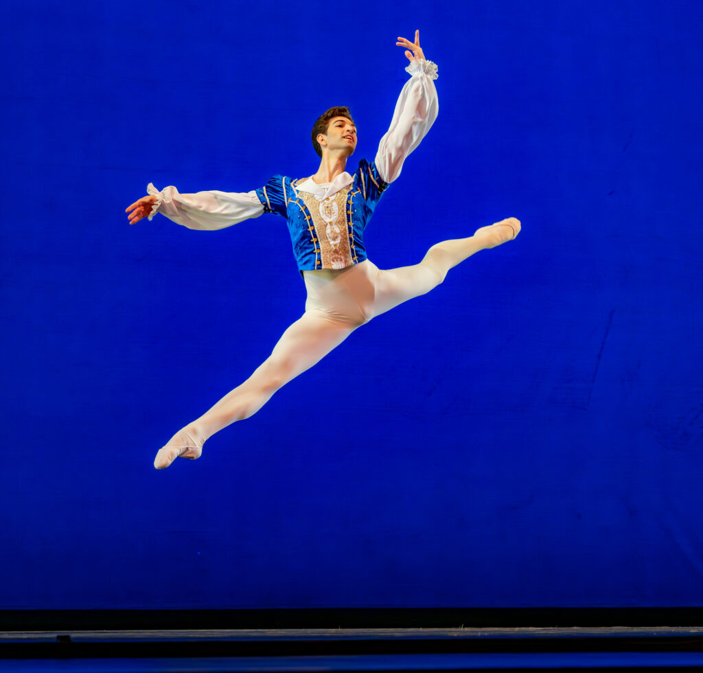 Yuval Cohen performs a sissone en avant, splitting his legs and holding his arms sin third position. He wears a blue tunic with white billowing sleeves, white tights and white ballet slippers and dances in front of a bright blue backdrop.