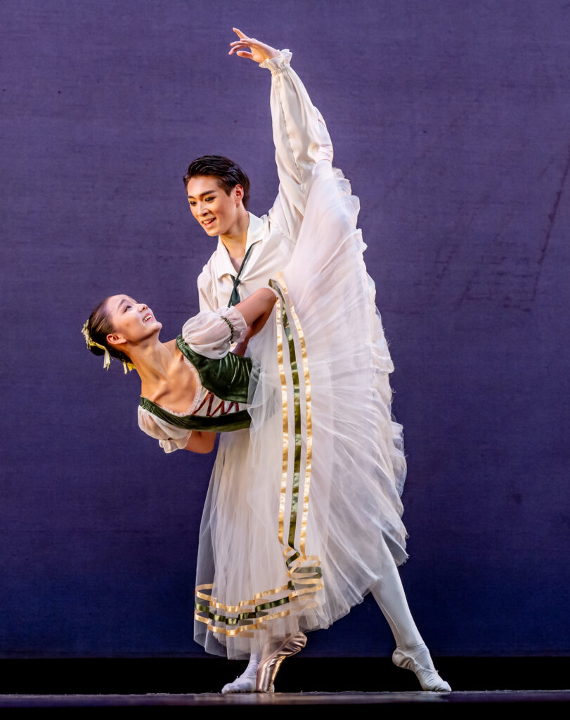 Mengxuan Yan and Zihan Kong perform a classical pas de deux onstage in front of a pruple backdrop. She does a penché in attitude with her arms bent and hands clasped behind her back and looks back at Zihan Kong, who holds her waist. She wears a white romantic tutu with a green peasant-style bodice, and he wears a white tunic and white tights and ballet slippers.
