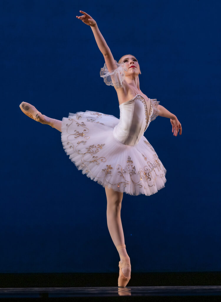Julie Joyner performs a pique attitude in croisé during a performance with her left lag in back. She raises her right arm and looks up toward it. She wears a white classical tutu, pink tights and pink pointe shoes and dances in front of a blue backdrop.