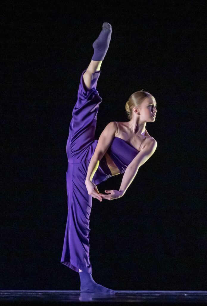 Isabella Howard does a high grnad battement with her right leg and leans her body over to the left, clasping her hands. She wears loose purple pants and a purple tube top and dances in front of a black backdrop.