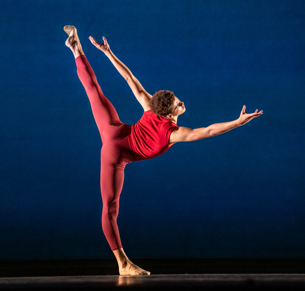 Corbin Holloway wears a red sleeveless top and red footless tights. His body faces the back of the stage and he does a penche with his left leg behind him, reaching his right arm out and stretching his left arm up and back.