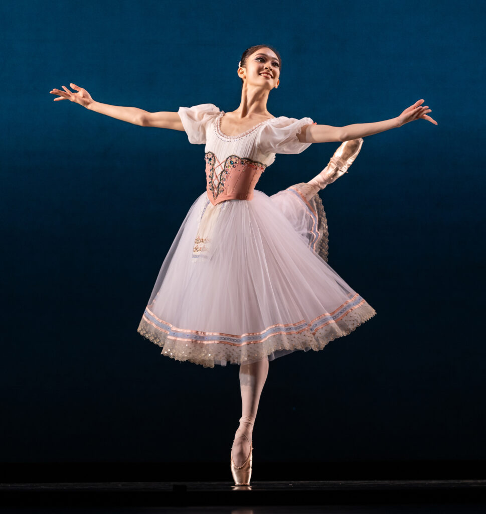 Yunju Lee does a piqué attitude croisé on pointe with her right leg back and her arms open in second with ehr palms facing up. She wears a light pink peasant costume with a knee-length tutu.