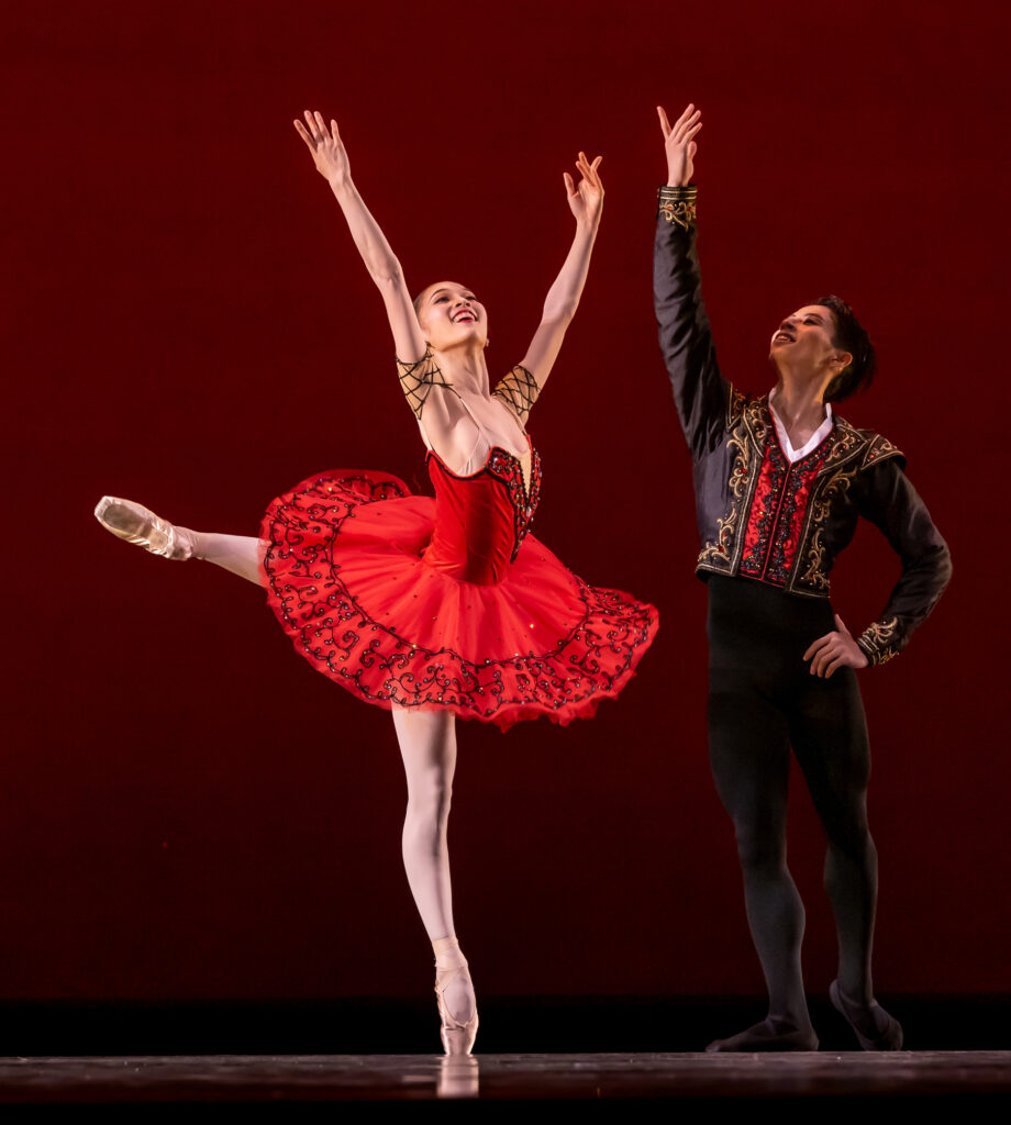Sayoko Taku and Ryo Sasaki dance the wedding pas de deux from the ballet Don Quixote. Toku wears a bright red classical tutu and poses ith her left leg in attitude croisé and her arms raised in a V shape, palms facing in. Sasaki stands back and to the right of her, his left hand on his hip and his right arm raised triumphantly. He wears a black a gold Spanish-style tunic and black tights.