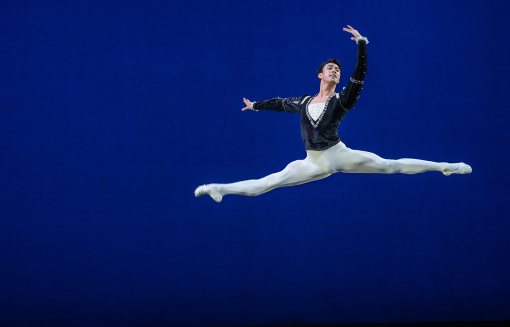 Munkjin Ulziijargal performs a grand jeté to the right and holds his arms in third position. He wears a blue velvet tunic, white tights and white ballet slippers and dances in front of a blue backdrop. He looks out to the audience with a proud smile.