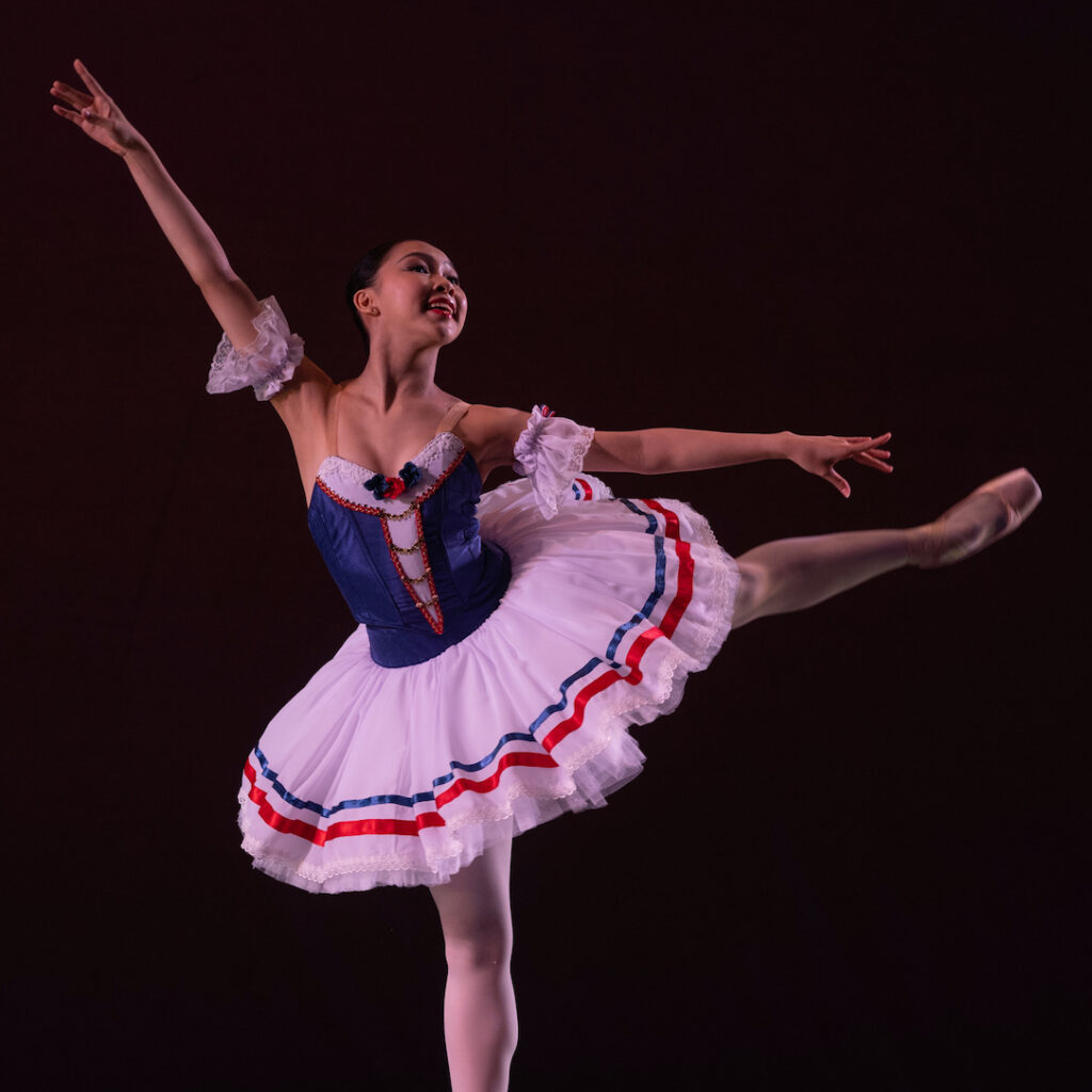 Nicole Liu does a first arabesque onstage during a performance with her left leg raised, She looks out to the audience with a big, spirited smile. She wears a costume with a blue bodice and white tutu with red and blue trimmings, pink tights and pink pointe shoes. She dances in front of a black backdrop.