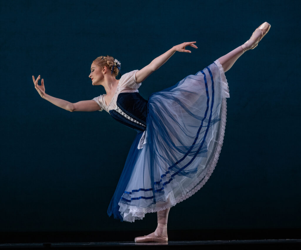 Grace Reid performs an arabesque elongé during a performance onstage, with her right leg raised and her right arm curved up. She smiles peacefully and looks down towards the floor. She wears a blue and white peasant dress costume, pink tights and pointe shoes.