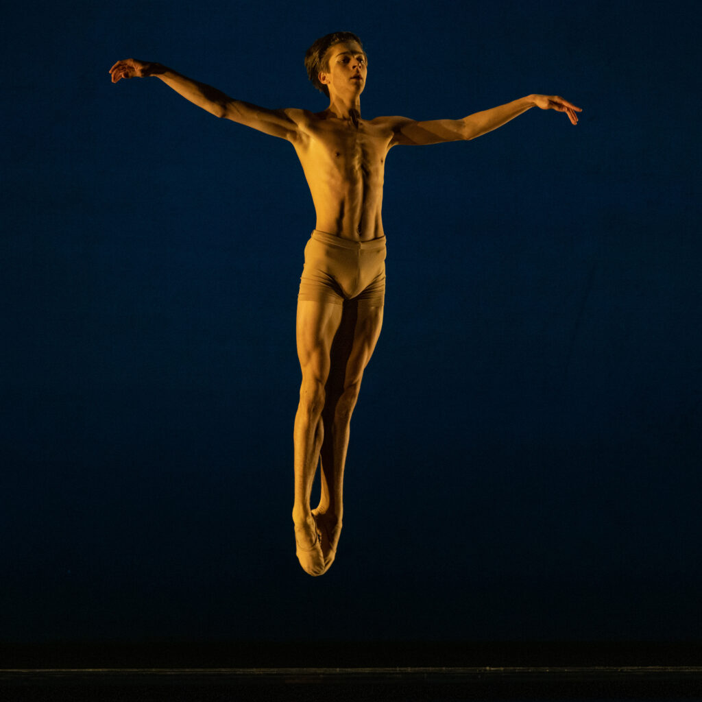 Ethan Maas jumps up into the air in an entrechat six, holding his arms out to the side, during a performance on a darkened stage. She wears short tan bike shorts and tan ballet slippers.