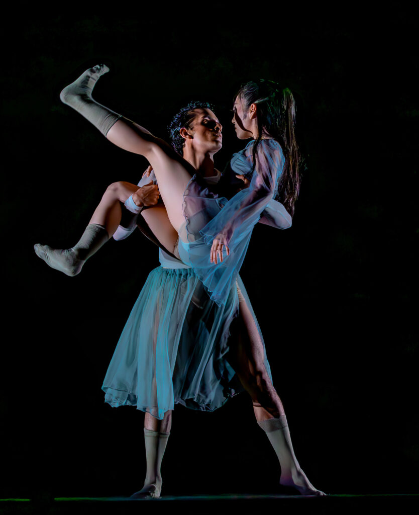On a drakened stage, Alejandro Gonzalez stands with his feet apart and holds Mayu Odaka in his arms. She kicks her left leg up slightly as her left arm hangs down, and the two dancers look intently at each other. Gonzalez wears a filmy turquoise skirt and green socks, while Odaka wears a filmy turquoise long-sleeved shirt and green socks.