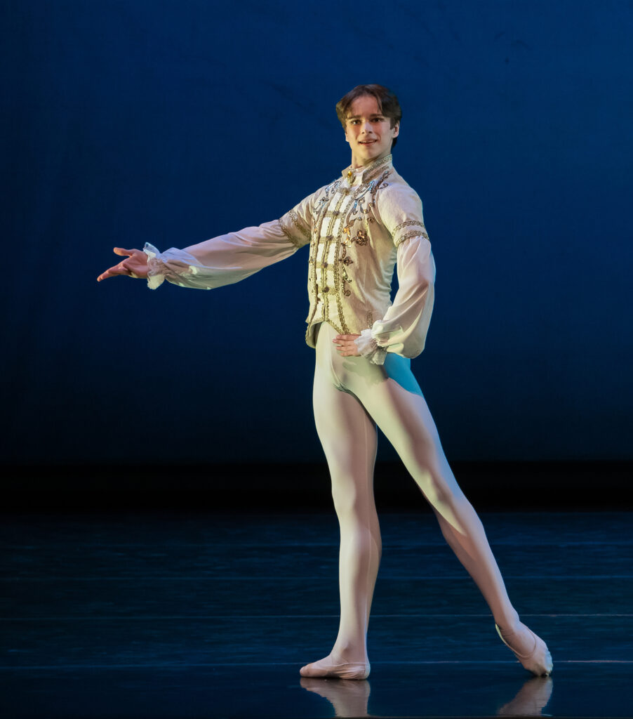 Alexei Orohovsky stands in tendu derriere in effacé during a performance. He stands on his right foot and extends his right arm out, palm up, and places his left hand on his hip as he looks out to the audience with a smile. He wears a princely white tunic with gold trim, white tights and white ballet slippers, and performs in front of a blue backdrop.