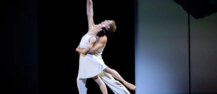 Amber Scott and Adam Bull dance a pas de deux onstage in front of abstract black and white sets. Bull holds her around the waist with his left arm as she does a low arabesque, pushing over her pointe shoe and thrusting her hips forward. They both left their right arms high, her hand draped over his forearm, and look up towards it. Scott wears a short white dress and Bull wears wide-legged white pants and a white tank top.
