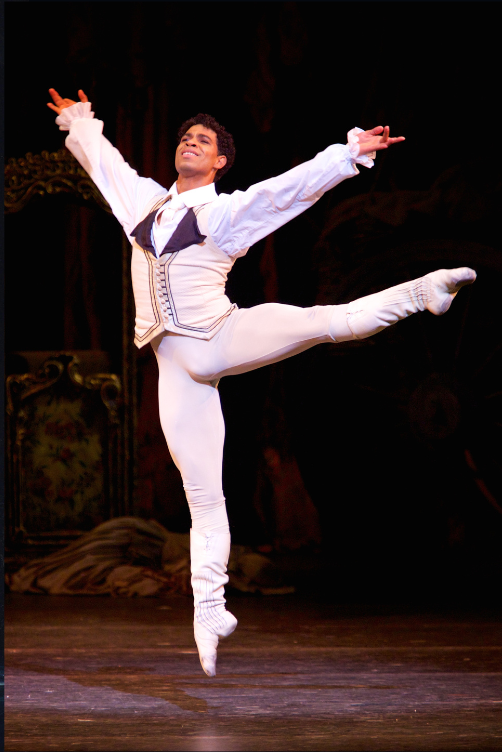 Carlos Acosta does a jubilant sauté with his left leg a la seconde at 90 degrees. He wears a white aristocrat-style costume and lifts his arms with confidence.