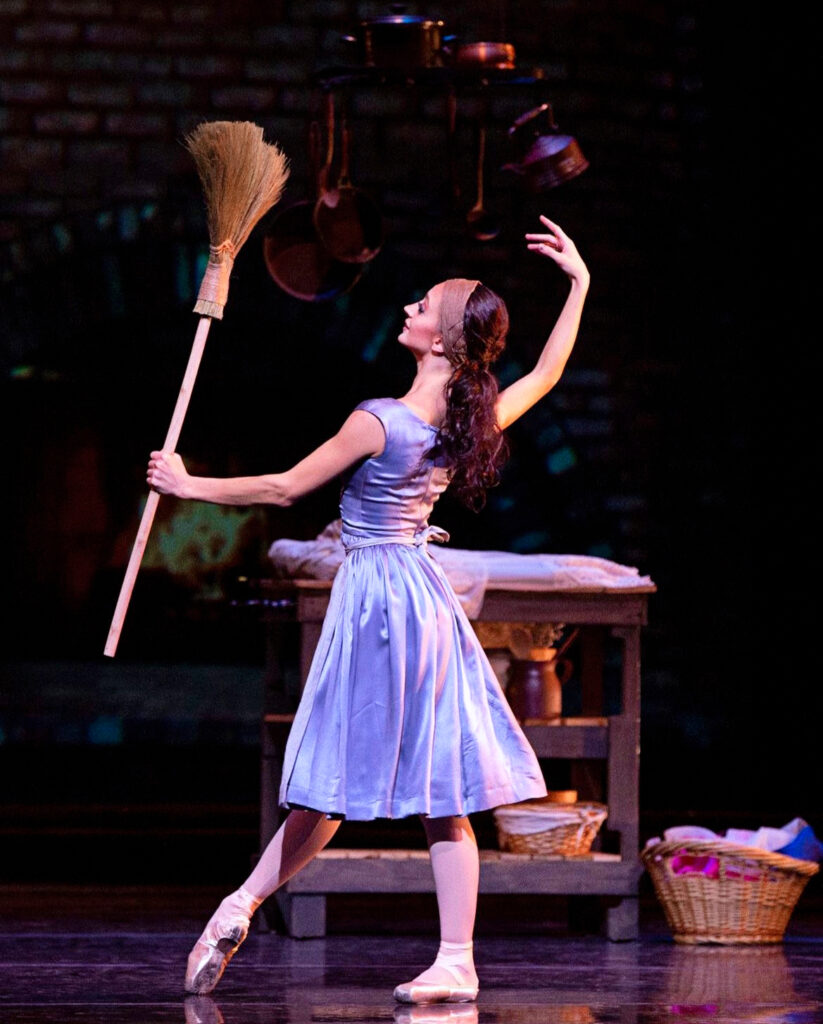 Buse Babadag performing "Cinderella." She is wearing a lavender dress, her right leg extended forward in tendu. She is holding a broom with her left hand and extending her right arm up.
