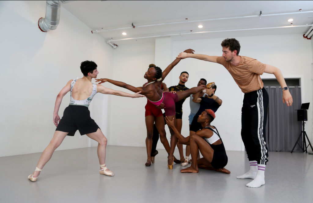 In a bright ballet studio, members of the Ballet22 Momentum program rehearse together. They all wear pointe shoes and hold on to each other in a contemporary tableau, Ashton Edwards in a pitched arabesque on center.