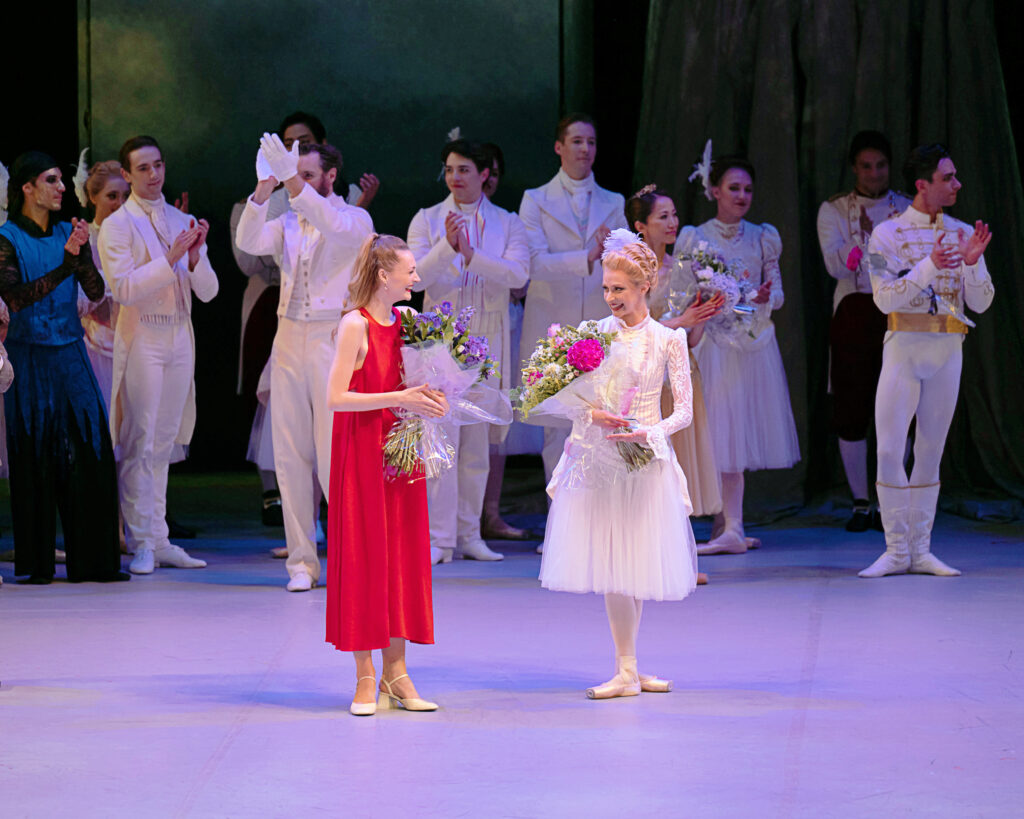 Breanna Foad and Paige Bestington stand together onstage in front of the English National Ballet company, in full costume, as they each hold a bouquet of flowers and smile.