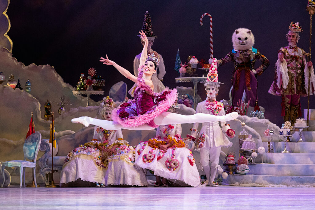 Beckanne Sisk—wearing a bright magenta tutu, large sparkly crown, pink tights and pointe shoes—does a large saut de chat towards stage right during a performance of Nutcracker. She dances in front the the Land of Sweets set as other costumed characters watch.