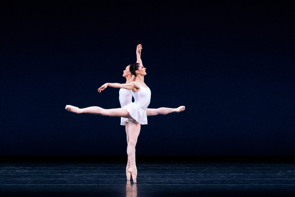 During a performance, Beckanne Sisk and Bridget Kuhns dance close together in arabesque, holding one arm up and holding each other's raised hand. Sisk is closer to the audience. Both dancers wear pink tights, pointe shoes, white leotards, and short white skirts.