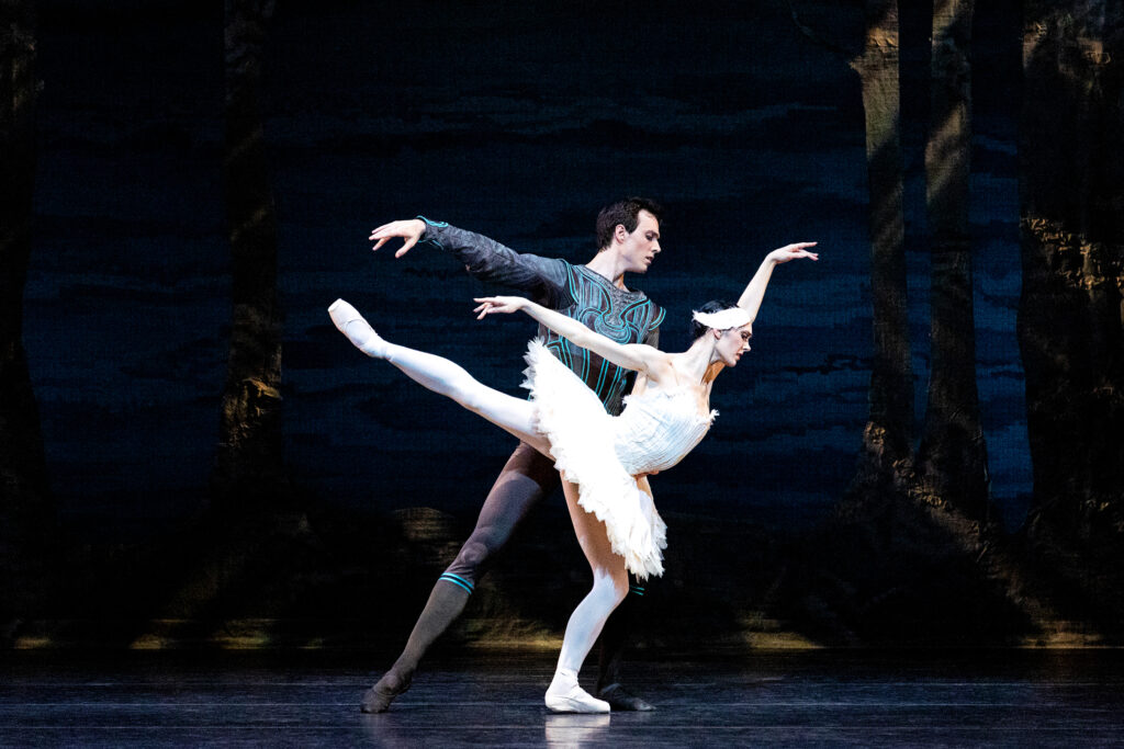 Beckanne Sisk and Chase O'COnnell perform a pas de deux from Swan Lake onstage. Sisk does an arabesque allongé towards stage left, while O'Connell stands behind her in tendu, extends his right arm out and holds her waist with his left hand. She wears a white tutu and feathered headpiece, pink tights and pointe shoes, while he wears a dark tunic and tights with blue trim. They perform in front of a lakeside backdrop.