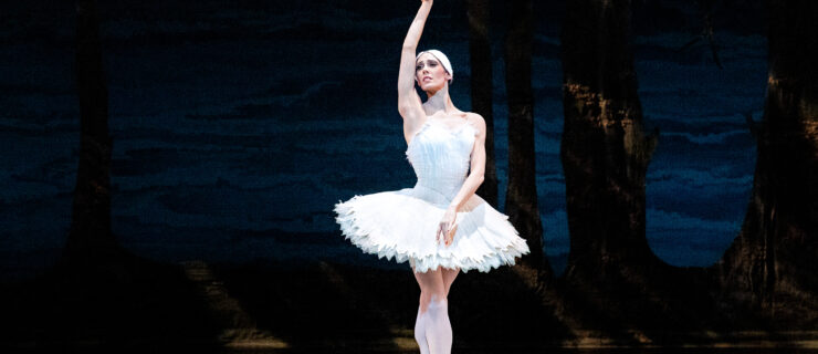 During a performance of Swan Lake, Backanne Sisk stands center stage in sus-sous on pointe, her right arm held high above her head, palm facing out, and her left arm low across her body. She turns her head towards her right arm. She wears a white tutu and feathered headpiece and dances in front of a dark backdrop showing a lakeside scene.