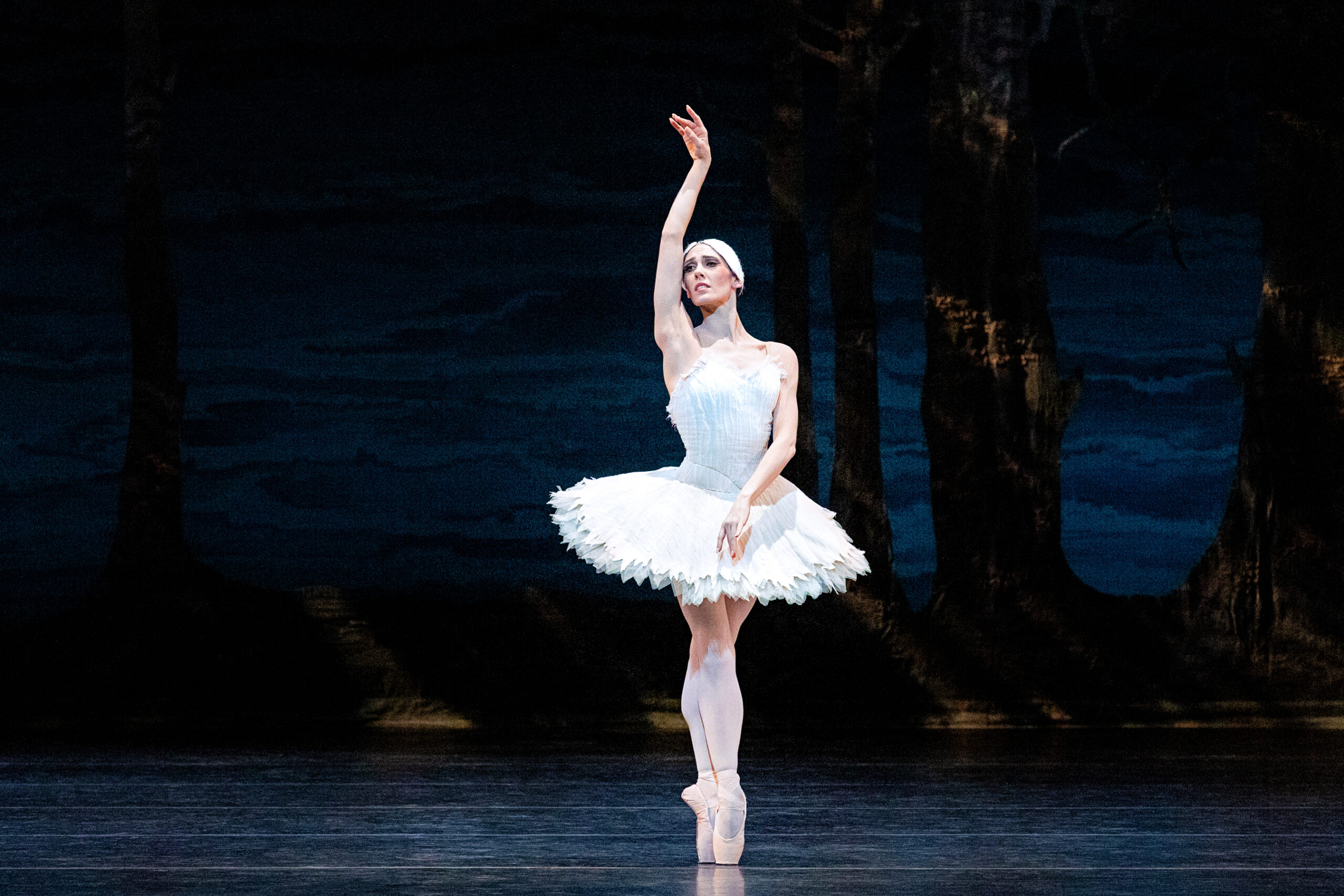 During a performance of Swan Lake, Backanne Sisk stands center stage in sus-sous on pointe, her right arm held high above her head, palm facing out, and her left arm low across her body. She turns her head towards her right arm. She wears a white tutu and feathered headpiece and dances in front of a dark backdrop showing a lakeside scene.