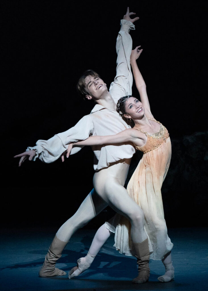 Cameron Catazaro and Nina Fernandes perform as Romeo and Juliet onstage, smiling joyously. Catazaro stands in a fourth position croisé closely behind Fernandes as she lunges in a tendu derriere. Both dancers open their amrs wide, with the backs of Fernandes' wrists touching Catazaro's arms. She wears a peach, empire waisted dress and pointe shoes, while he wears a white top with billowy long sleeves, off white tights and brown, calf-length boots.