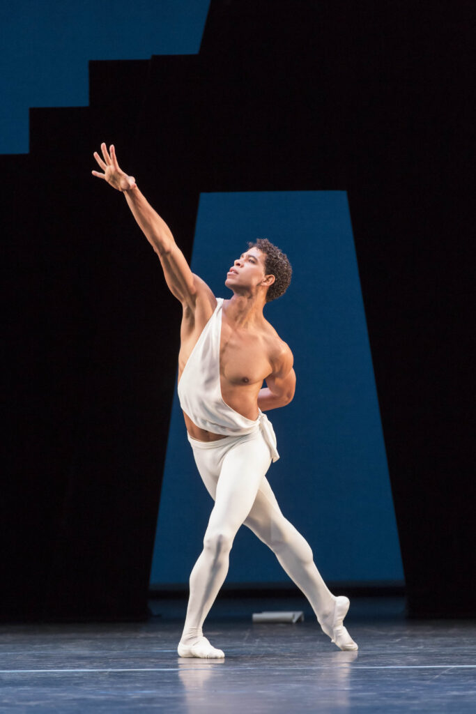 Carlos Acosta performs in George Balanchine's "Apollo," wearing a white sash and white tights. He lunges forward in croise, reaching his right hand upward on the diagonal and bending his left arm behind his back.
