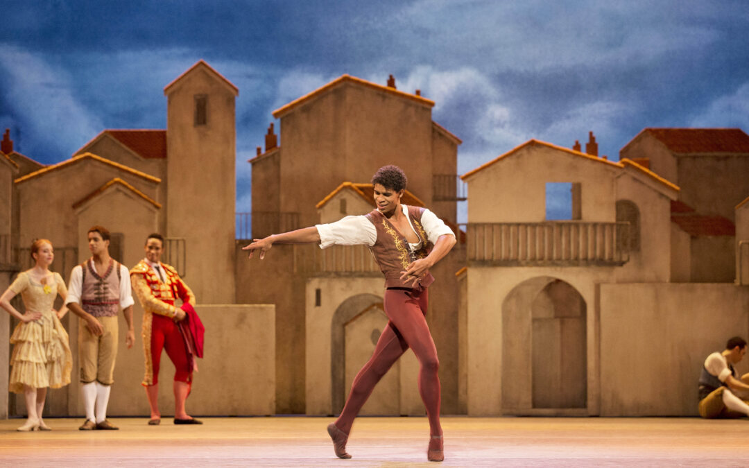 Carlos Acosta performs in Don Quixote on the Royal Opera House stage. He wears a tunic and tan vest with rust-colored tights and poses passionately in a tight lunge on forced arch, looking back at his extended right arm.