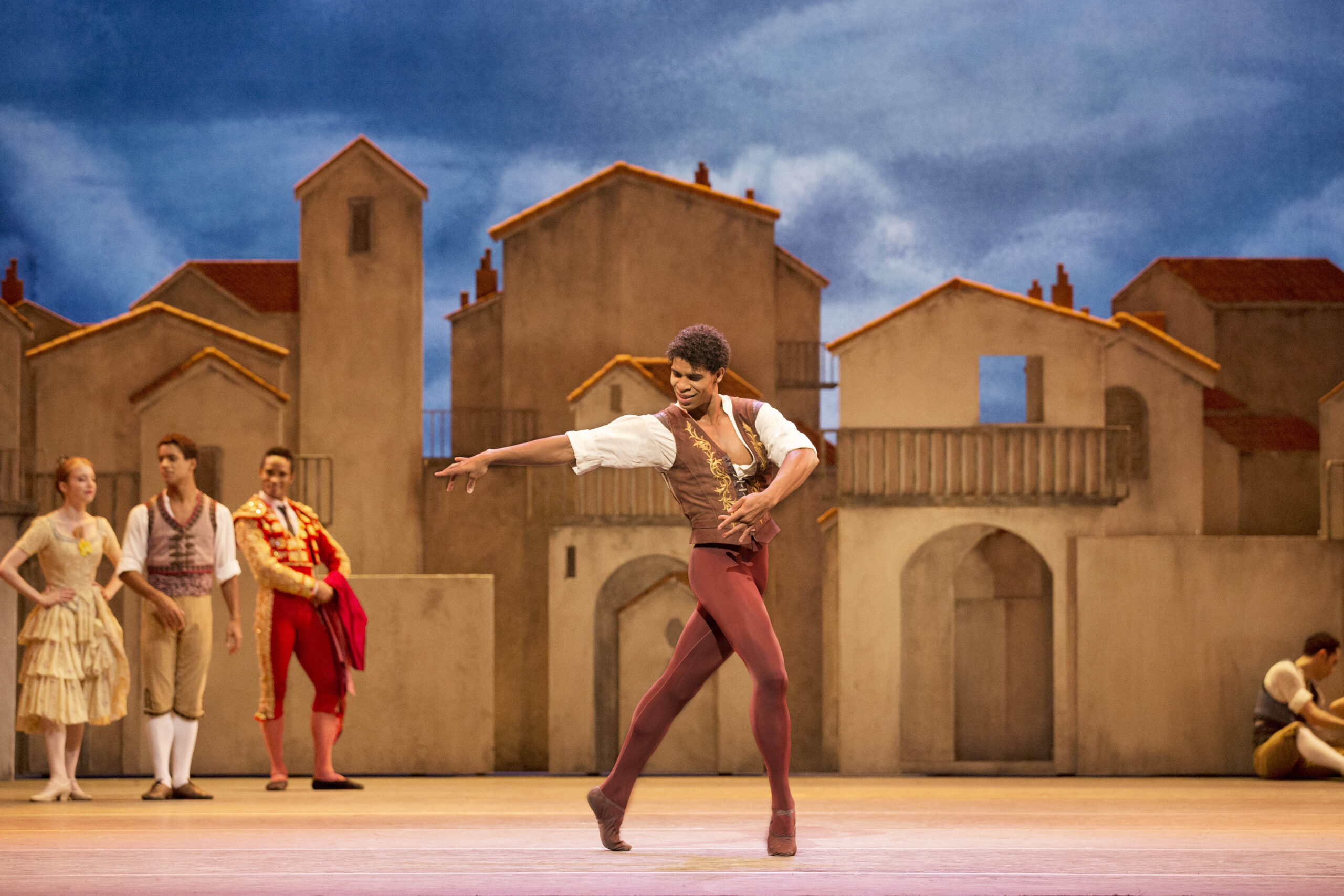 Carlos Acosta performs in Don Quixote on the Royal Opera House stage. He wears a tunic and tan vest with rust-colored tights and poses passionately in a tight lunge on forced arch, looking back at his extended right arm.
