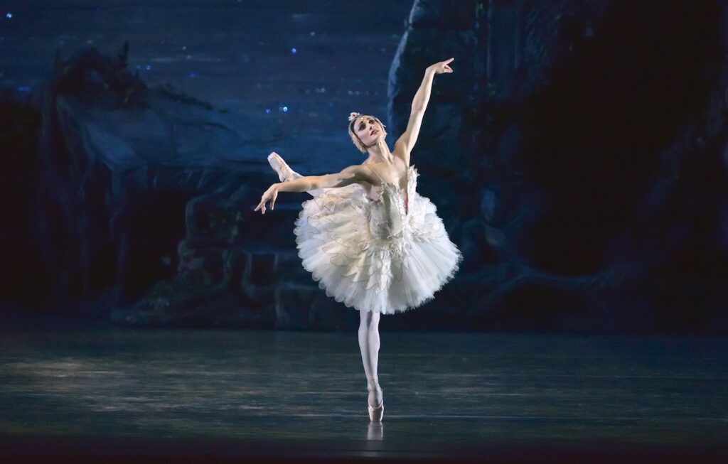 Sarah Lane performing in Swan Lake. She poses in a dramatic arabesque on face, arms lifting.