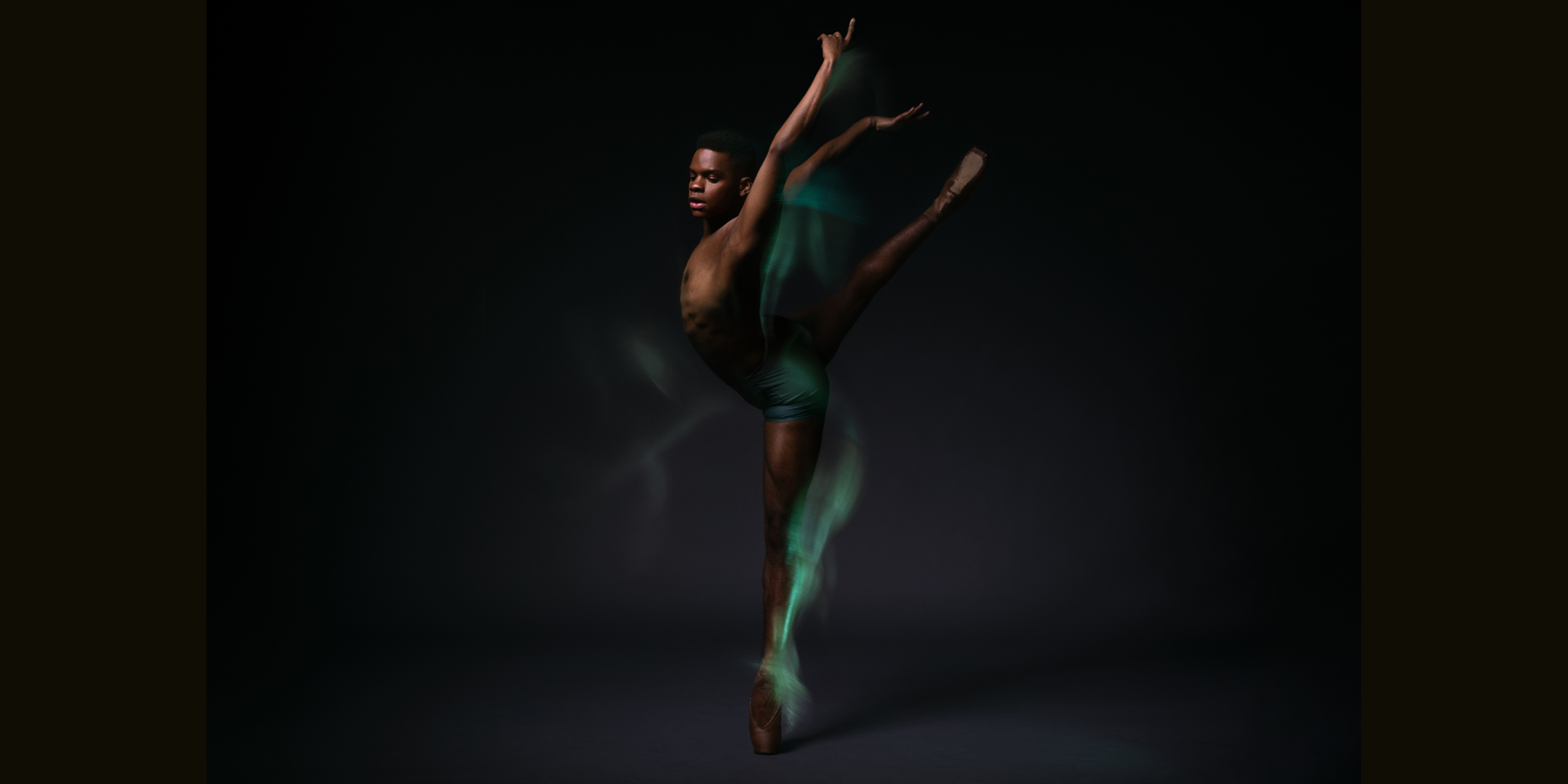 Daniel R. Durrett poses in a dramatic attitude croise derriere on pointe, in front of a pitch black background. Green and purple wisps of color trace their bottom leg and arms.
