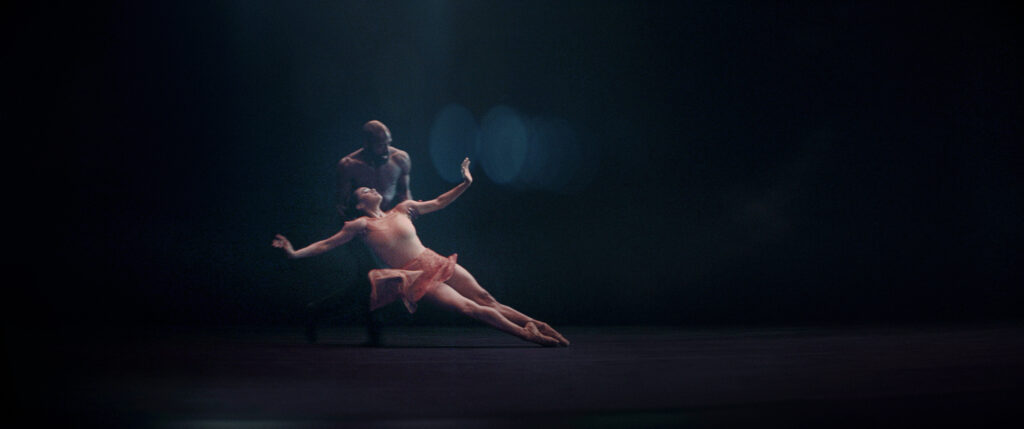 Misty Copeland and Babatunji Johnson dance a contemporary pas de deux against a dark background. She is wearing a peach-colored leotard and skirt