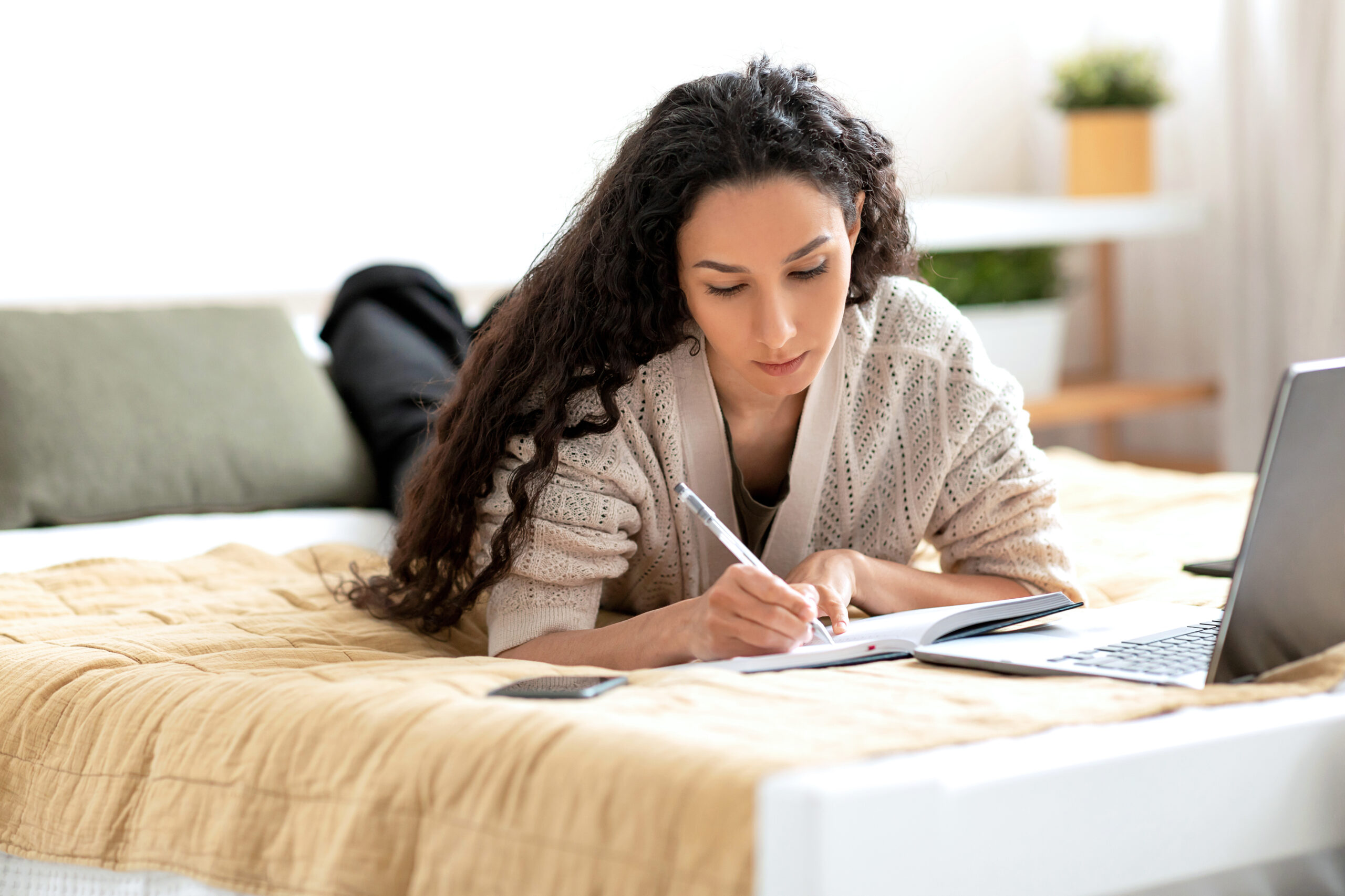 A young woman with long, curly dark hair lies on a bed, using a laptop and writing in a notebook.