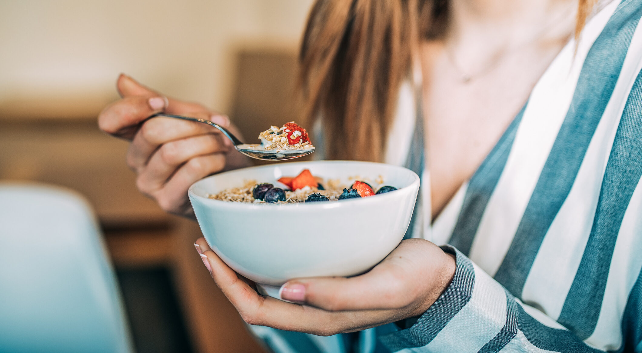 A cropped photo of a woman, shown close up, eating a bowl of oat and fruit cereal for breakfast.