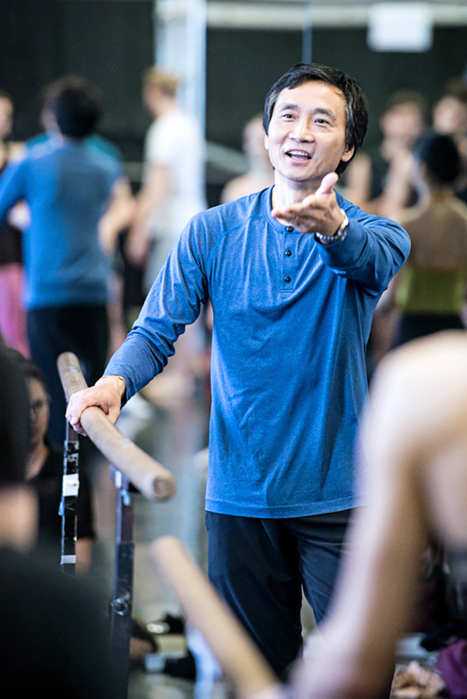 Li Cunxin leads company class at Queensland Ballet. He wears a longsleeve blue shirt and smiles as he lifts his left arm forward, palm upward, with his other hand resting on the bar.