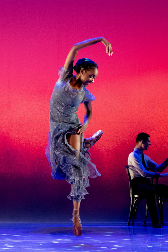 Ashley Simpson, wearing a long purple dress with a slit up the left side of the skirt, stands on pointe and kicks her right foot back. She beds her body over to the left, her right arm high, and looks down towards her back foot.