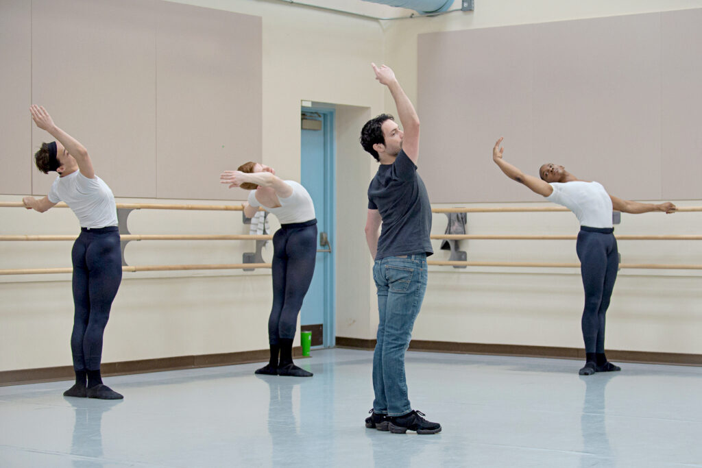 A male ballet teacher wearing jeans and a black T-shirt stands in first position in the center of a studio and does a cambré back. Three male students in white T-shirts, black tights and black ballet slippers stand him at the barre behind him, doing the same step.