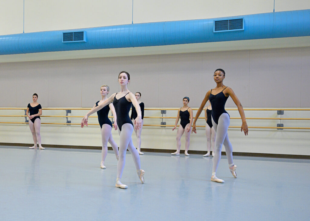 Three female ballet students stand en face in tendu derriere with their right leg in back. They hold their arms in demi-seconde position. They wear black leotards, pink tights and pink pointe shoes. Four other ballet dancers in the same dancewear stand behind them, observing.