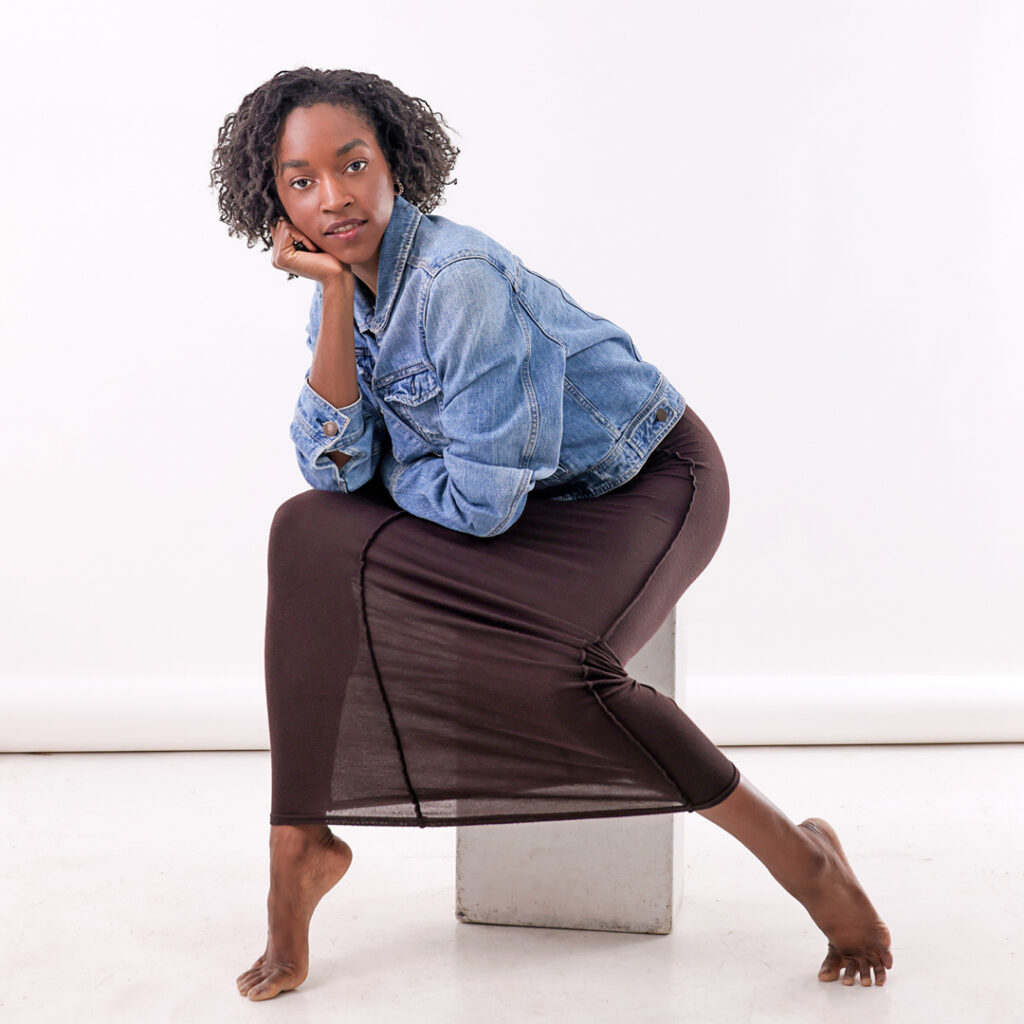 Ashley Simpson. wearing a jean jacket and black maxidress, sits on an apple box in front of a white backdrop. She turns her body slightly to the right corner, extends her left leg out long, props her right elbow on her knee and rests her chin in her right hand. She smiles sweetly towards the camera.