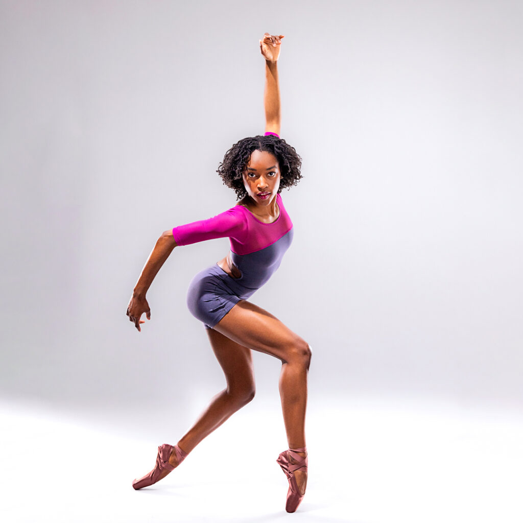 Ashley Simpson is shown on pointe facing stage left with her legs in a wide, parallel stance and her knees bent. She smiles and twists her upper body to face front, opening her right arm out and lifting her left arm straight up behind her head. She wears a two-tone fuchsia and gray biketard with three-quarter sleeves. She poses in front of a light gray backdrop.