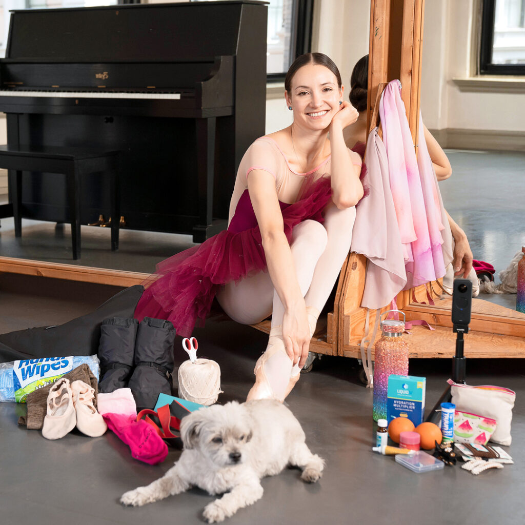 Christine Shevchenko poses with her dance-bag items and her dog, Rey, in an ABT studio. She is seated and smiling, with her face resting on her left hand.