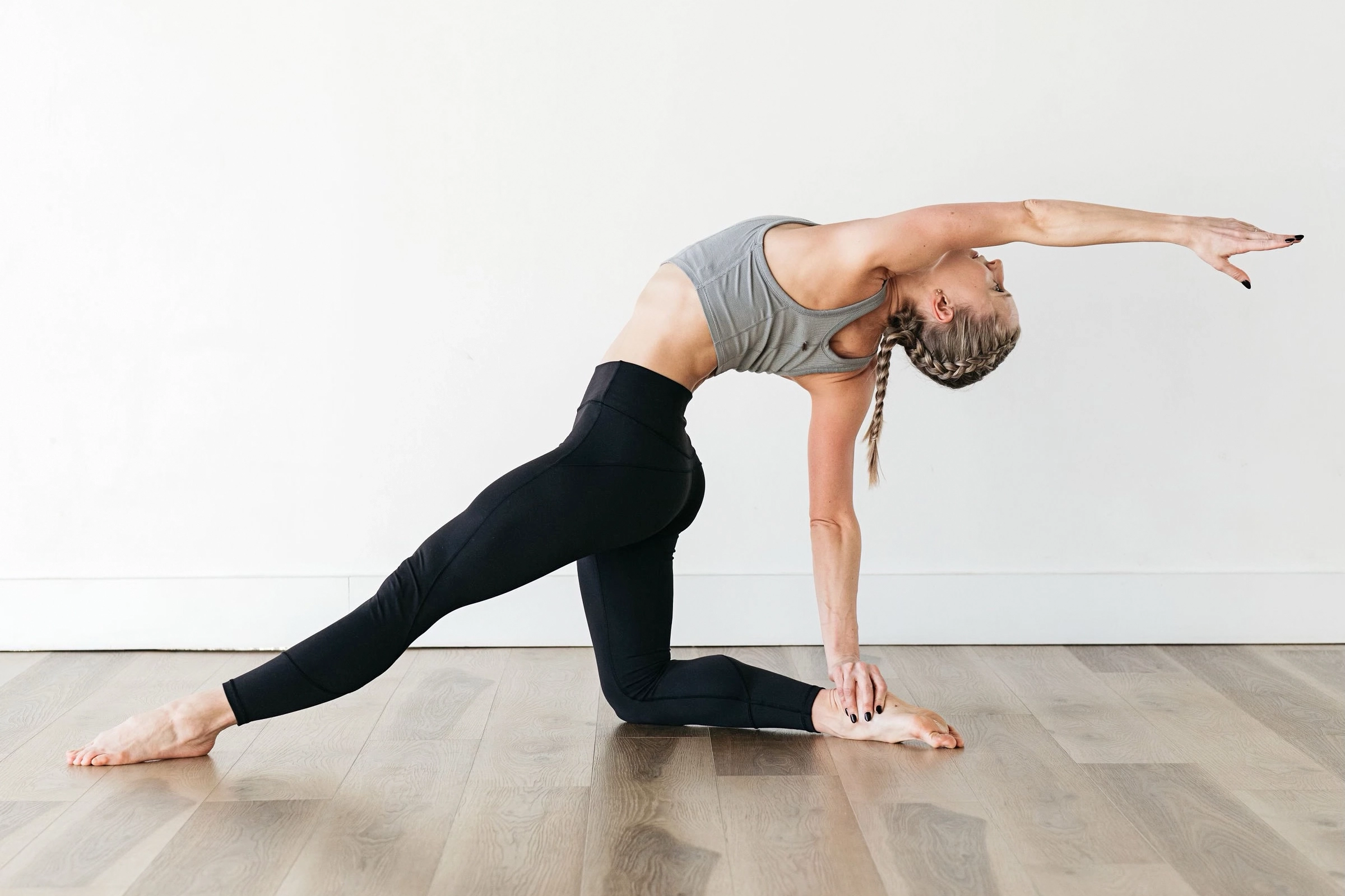 Leigh Anne Albrechta wears a gray sports bra and black leggings and does a yoga pose on a wooden floor. She balances on her right know and extends her left leg out while holding her right heel with her right hand and reaching back with her left arm and upper body.