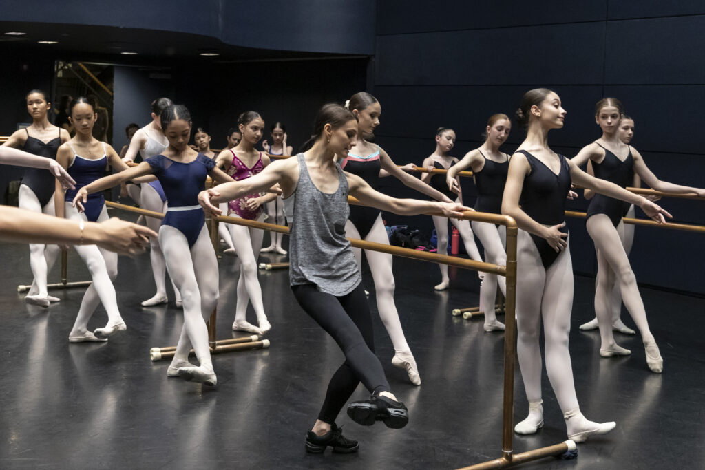 In a black box studio, Sarah Lane demonstrates a degage combination to a class of young dance students. She holds on to the barre gently and shows a fondu degage devant with her foot flexed.