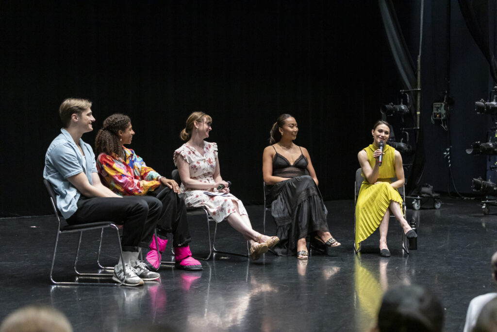In a large black box studio space, Cameron Catazaro, Adji Cissoko, Amy Brandt, Candy Tong, and Sarah Lane sit in a shallow semi-circle for a panel discussion. Lane holds the microphone and speaks; the other four panelists listen and smile.
